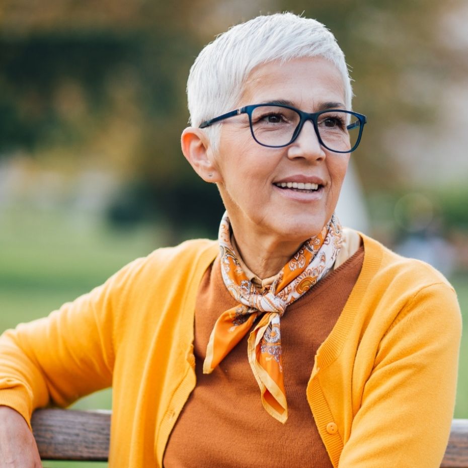 Frau im Gleitsichtalter mit Brille sitzt auf einer Parkbank im Grünen.